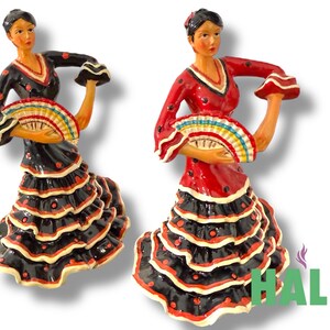 May include: Two ceramic figurines of flamenco dancers. One dancer wears a black dress with red polka dots and a ruffled skirt. The other dancer wears a red dress with black polka dots and a ruffled skirt. Both hold a colorful fan.