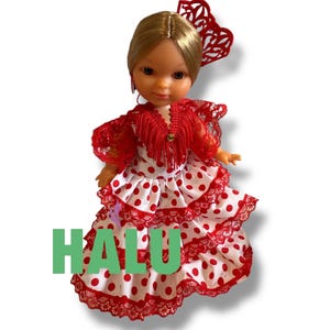 May include: A doll wearing a traditional Spanish flamenco dress. The dress is white with red polka dots and red lace trim. The doll has blonde hair and a red hair accessory. The word "HALU" is printed in green.