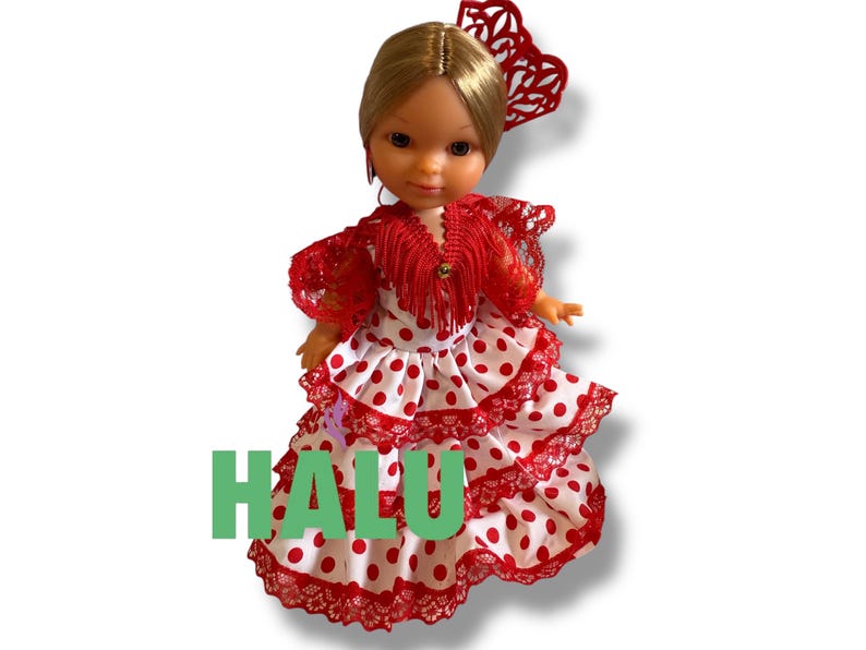 May include: A doll wearing a traditional Spanish flamenco dress. The dress is white with red polka dots and red lace trim. The doll has blonde hair and a red hair accessory. The word "HALU" is printed in green.