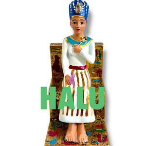 May include: A figurine of an Egyptian pharaoh sitting on a throne. The pharaoh is wearing a white robe with gold stripes, a red sash, and a blue and gold crown. The throne is decorated with gold and hieroglyphics. The word "HALU" is written in green letters below the throne.