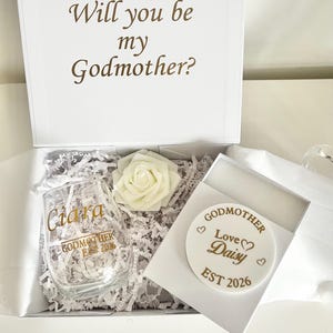 Könnte beinhalten: Eine weiße Geschenkbox mit dem Text "Will you be my Godmother?" in brauner Schrift. Im Inneren ein stieloses Weinglas mit goldfarbenen Buchstaben, eine weiße Rose und ein runder Untersetzer mit der Aufschrift "Godmother Love Daisy EST 2026".