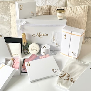 May include: White gift boxes with gold lettering and ring designs. The boxes are numbered 1 through 7. The boxes contain a variety of items, including a bottle of champagne, a candle, a glass, and a robe. The boxes are arranged on a white bedspread.