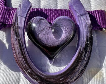 May include: A purple horseshoe-shaped pendant with a heart-shaped centre. The heart is filled with dark hair, and the horseshoe is accented with dark hair. The pendant is attached to a purple strap.