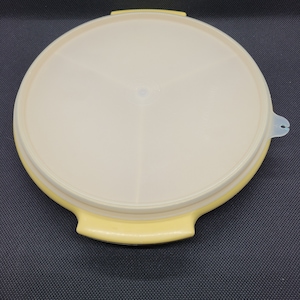 Op de afbeelding: Ronde serveerschaal van geel plastic met een doorzichtig deksel. Het deksel heeft een klein handvat aan de zijkant.