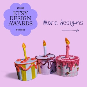 Pode incluir: Três objetos cilíndricos em forma de bolo com velas sobre um fundo roxo claro. O texto diz 2026 ETSY DESIGN AWARDS Finalist e More designs com uma seta. Os bolos apresentam listras amarelas, laços roxos e padrões xadrez cor-de-rosa.