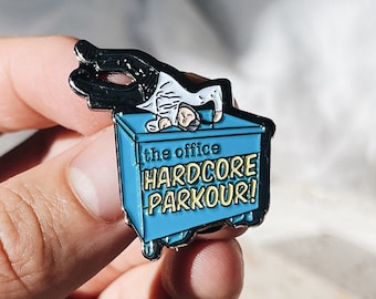 Könnte beinhalten: Ein blauer Emaille-Pin mit einer Figur aus "The Office", die Parkour auf einem blauen Müllcontainer macht. Der Pin trägt den Text "the office HARDCORE PARKOUR!" in Gelb und Weiß.