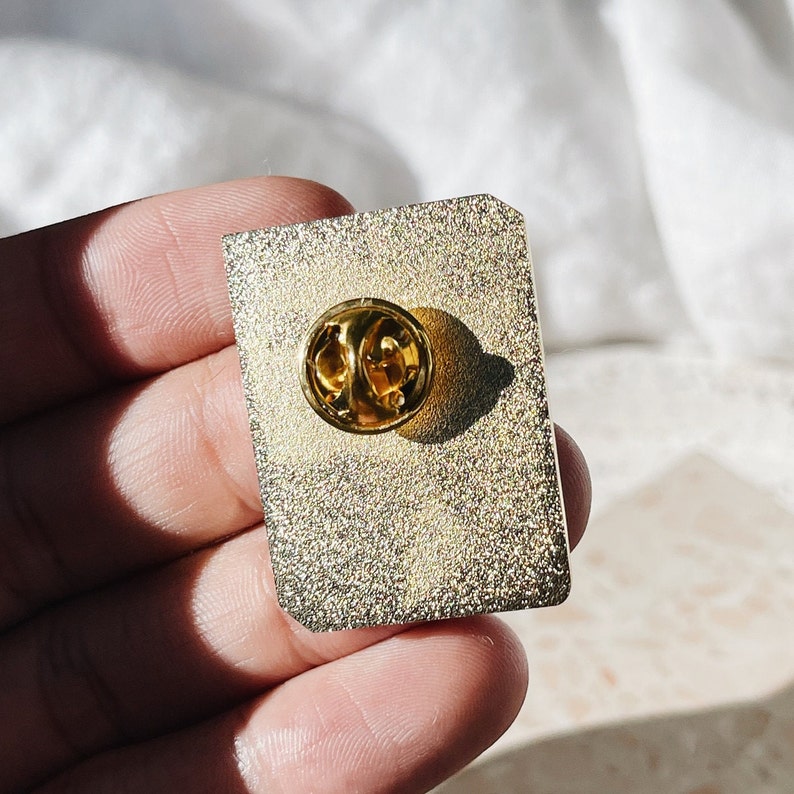 May include: A rectangular gold-colored enamel pin with a gold-colored butterfly clutch. The pin has a textured surface and a gold border. The pin is designed to be attached to clothing or accessories.