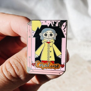 May include: Enamel pin featuring a character from the movie "Coraline." The pin has a pink border and a black background with a yellow moon and stars. The character wears a yellow coat and has button eyes. The word "Coraline" is written in cursive.