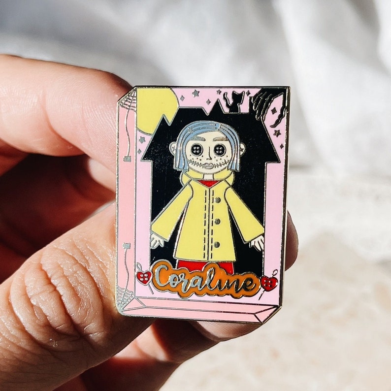 May include: Enamel pin featuring a character from the movie "Coraline." The pin has a pink border and a black background with a yellow moon and stars. The character wears a yellow coat and has button eyes. The word "Coraline" is written in cursive.