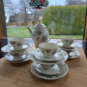 May include: A white tea set with black floral designs. The set includes a teapot, four cups, and four saucers. The cups and saucers are stacked on top of each other.