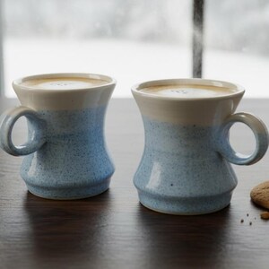 Peut inclure: Deux tasses en céramique bleues et blanches remplies d'une boisson crémeuse sont posées sur une table en bois foncé. Un biscuit rond brun doré est posé à côté des tasses. Les tasses ont une forme évasée et des anses arrondies.