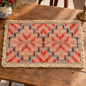 May include: A rectangular woven placemat featuring a geometric pattern in shades of red, light pink, and blue on a natural tan background. The placemat is finished with a decorative fringe along all four edges, creating a textured border.
