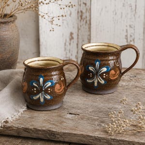 Peut inclure: Deux tasses en céramique arrondies avec un vernis brun moucheté, ornées d'un motif floral peint à la main en bleu et blanc avec des accents en spirale orange. Chaque tasse possède une large anse et un intérieur crème, avec une finition brillante.