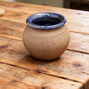 Puede incluir: Una olla redonda de cerámica hecha a mano, con un exterior beige texturizado y un borde azul oscuro. La olla está sobre una mesa de madera rústica. El fondo muestra una cocina con encimeras de madera.