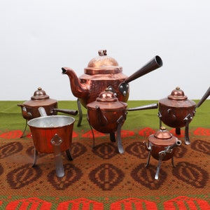 Könnte beinhalten: Ein Set aus fünf Kupferteekannen mit Henkeln und Beinen. Die Teekannen sind auf einer grün-braunen gemusterten Tischdecke angeordnet.