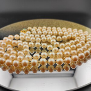 May include: A strand of cream-colored pearls on a reflective surface. The pearls are strung together with a gold clasp.