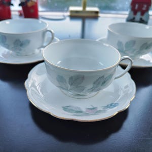 Puede incluir: Tres tazas de té blancas con un delicado patrón floral verde y ribete dorado. Cada taza de té está sobre un platillo a juego con un borde festoneado.