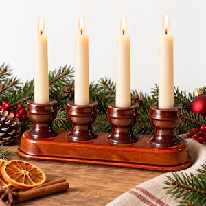 Pode incluir: Um castiçal de cerâmica castanha com quatro suportes circulares dispostos em fila, cada um contendo uma vela cónica longa e fina de cor branco-sujo. As velas estão acesas e emitem uma luz quente. A base é retangular com cantos arredondados.