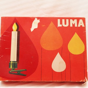 Puede incluir: Una caja roja con la palabra "LUMA" impresa en blanco. La caja presenta una vela blanca con una llama amarilla y un clip verde. El fondo de la caja tiene tres formas de gota de color amarillo, naranja y blanco.