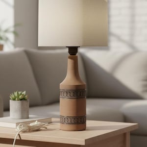 Puede incluir: Una lámpara de mesa de cerámica con una pantalla de tela beige. La base de la lámpara es de color marrón claro con un patrón decorativo negro. La lámpara está sobre una mesa auxiliar de madera, con una pequeña planta y un sofá en el fondo.