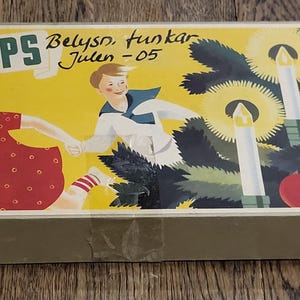 Könnte beinhalten: Vintage Philips Weihnachtslichtbox. Die Box hat einen gelben Hintergrund mit Illustrationen von Kindern, Kerzen und Äpfeln. Der Text "PHILIPS" ist deutlich sichtbar. Die Box ist hellbeige.