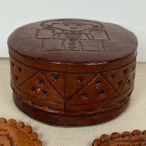 May include: A round, brown leather box with a lid featuring an embossed figure. The sides of the box are decorated with star and triangle patterns. The box is a decorative storage item.