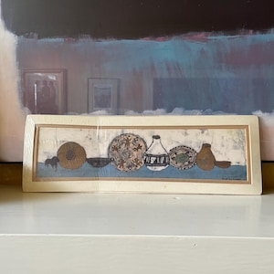 May include: A framed print featuring a collection of pottery. The artwork depicts various vessels and bowls in earthy tones of brown, beige, and blue. The print is encased in a cream-colored rectangular frame, resting on a white surface.