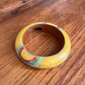 May include: A yellow wooden bangle bracelet with a painted design. The bracelet has a yellow exterior with a painted design of red, blue, and green stripes. The interior is a dark brown color.