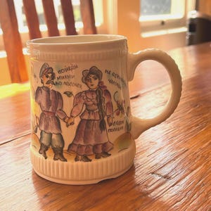 May include: Cream-colored ceramic mug with a handle. The mug features a hand-painted design of a couple in traditional clothing, holding hands. Russian text is visible. The mug has a ribbed base and a curved handle.