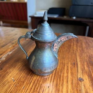 May include: A small, antique metal coffee pot with a pointed lid and a curved spout. The pot has a tarnished, aged appearance with shades of copper and green. The handle is small and curved, and the pot sits on a wooden surface.