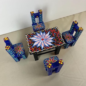 May include: A miniature wooden table and chairs set. The table has a black base and a top decorated with a large floral design in red, white, and blue. The chairs are blue with floral patterns. The set is painted with bright colors.