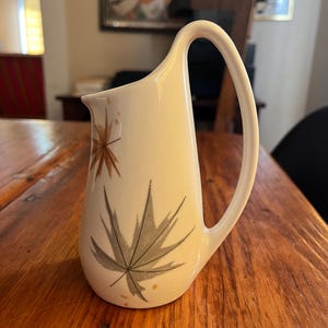 May include: Cream-colored ceramic pitcher with a large, curved handle. The pitcher features a design of stylized gray and brown leaves. The pitcher is likely vintage and suitable for serving beverages.