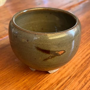 May include: A white ceramic mug with a black handle and the text "But First, Coffee" in a bold, black font. The mug is set against a blurred background, suggesting a cozy setting.