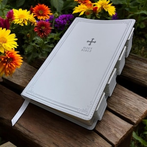 May include: White leather-bound Holy Bible with a silver cross and embossed lettering on the cover. The Bible is closed and lying on a wooden surface.