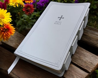 May include: White leather-bound Holy Bible with a silver cross and embossed lettering on the cover. The Bible is closed and lying on a wooden surface.