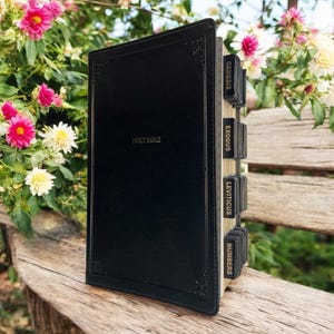 May include: A black leather bound Holy Bible with gold lettering on the front cover. The spine has gold tabs with the names of the first four books of the Old Testament: Genesis, Exodus, Leviticus, and Numbers.