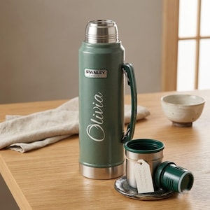 May include: A sage green Stanley vacuum insulated thermos with a silver metallic cap and base. The name Olivia is printed in white script on the side. A matching green and silver cup sits next to it with a small white paper tag tied with twine.