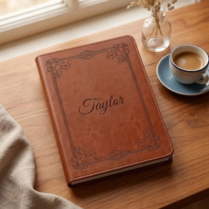 Op de afbeelding: Een bruin notitieboek in lederlook met een donker gegraveerde rand met decoratieve hoekversieringen en de naam Taylor in het midden in een sierlijk lettertype. Het rechthoekige boek ligt op een houten oppervlak naast een koffiekopje.