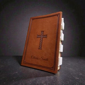 May include: A brown leather-bound Bible with a cross design and the name "Olivia Smith" embossed on the cover. The book has white tabbed pages with text labels. The Bible is set against a dark background.