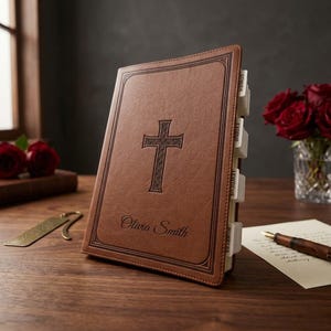 May include: A brown leather-bound Bible with a cross design and the name "Olivia Smith" embossed on the cover. The book has labeled page dividers and is displayed on a wooden surface with a bookmark and a pen.
