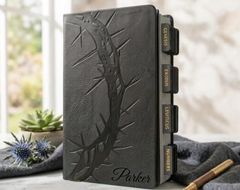 May include: A black leather-textured Bible featuring an embossed crown of thorns design on the front cover. The name Parker is printed in script at the bottom. Black index tabs with gold text label the books Genesis, Exodus, Leviticus, and Numbers.