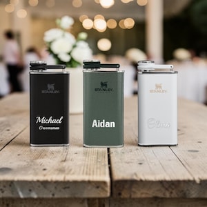 May include: Three Stanley flasks in black, green, and white, each with a personalized name. The black flask says "Michael Groomsman", the green flask says "Aidan", and the white flask says "Olivia". The flasks are on a wooden table.