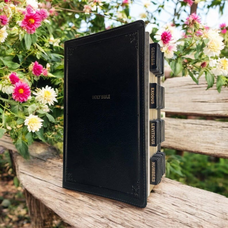 May include: A black leather bound Holy Bible with gold lettering on the front cover. The spine has gold tabs with the names of the first four books of the Old Testament: Genesis, Exodus, Leviticus, and Numbers.