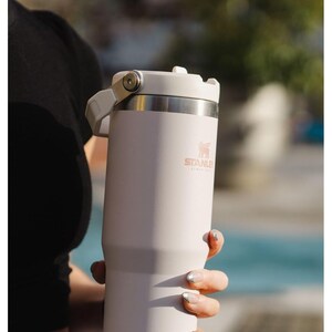 May include: A white stainless steel travel mug with a black handle and a rose gold Stanley logo. The mug has a lid with a flip top.