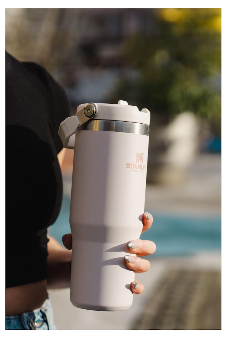 May include: A white stainless steel travel mug with a black handle and a rose gold Stanley logo. The mug has a lid with a flip top.