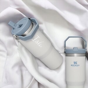 May include: Two stainless steel water bottles with blue lids and handles. The bottle on the left has the name "Isla" engraved on it. The bottle on the right has the Stanley logo and the text "Since 1913".