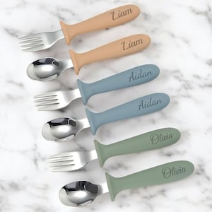 May include: Set of six personalized baby spoons and forks with blue, green, and tan handles. The handles are engraved with the names Liam, Aidan, and Olivia.