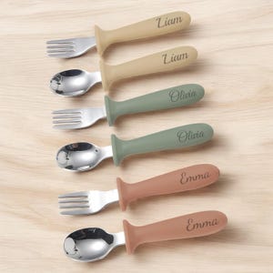 May include: Set of six personalized baby spoons and forks with silicone handles. The handles are in shades of brown, green, and beige. Each handle has a name engraved on it: Liam, Olivia, and Emma.