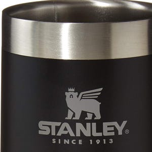 May include: Black insulated tumbler with a silver rim. The Stanley logo, featuring a bear with wings and a crown, is printed in silver. The text "STANLEY" and "SINCE 1913" are also visible.