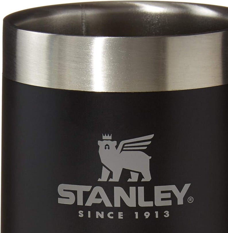 May include: Black insulated tumbler with a silver rim. The Stanley logo, featuring a bear with wings and a crown, is printed in silver. The text "STANLEY" and "SINCE 1913" are also visible.